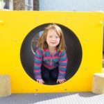 A young girl in a striped jacket crouches inside a yellow circular playground tunnel outdoors, smiling at the camera.