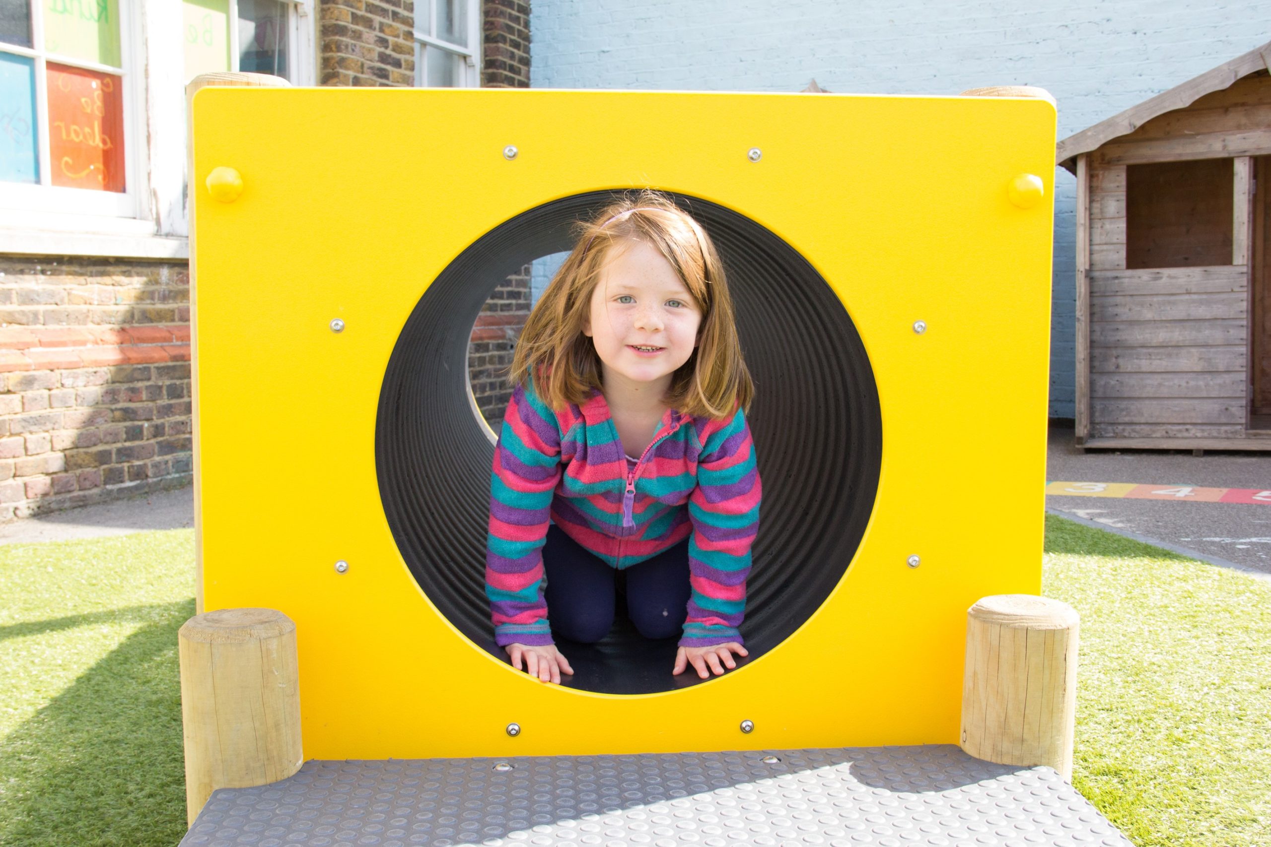 A young girl in a striped jacket crouches inside a yellow circular playground tunnel outdoors, smiling at the camera.