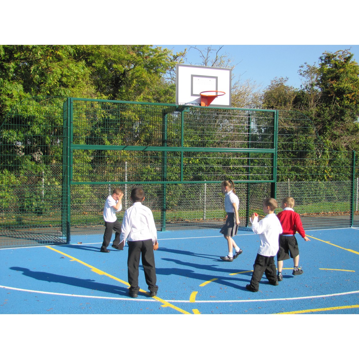 Five children play basketball on an outdoor court with a blue surface and a green metal fence, surrounded by trees.