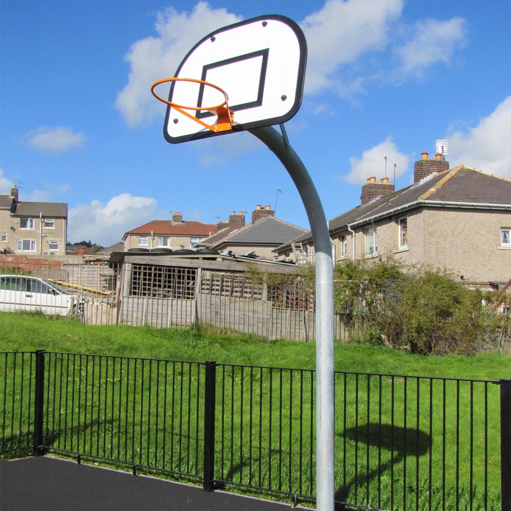 Outdoor basketball hoop with a white backboard and orange rim, installed on a tarmac court beside a grassy area and residential houses under a partly cloudy sky.