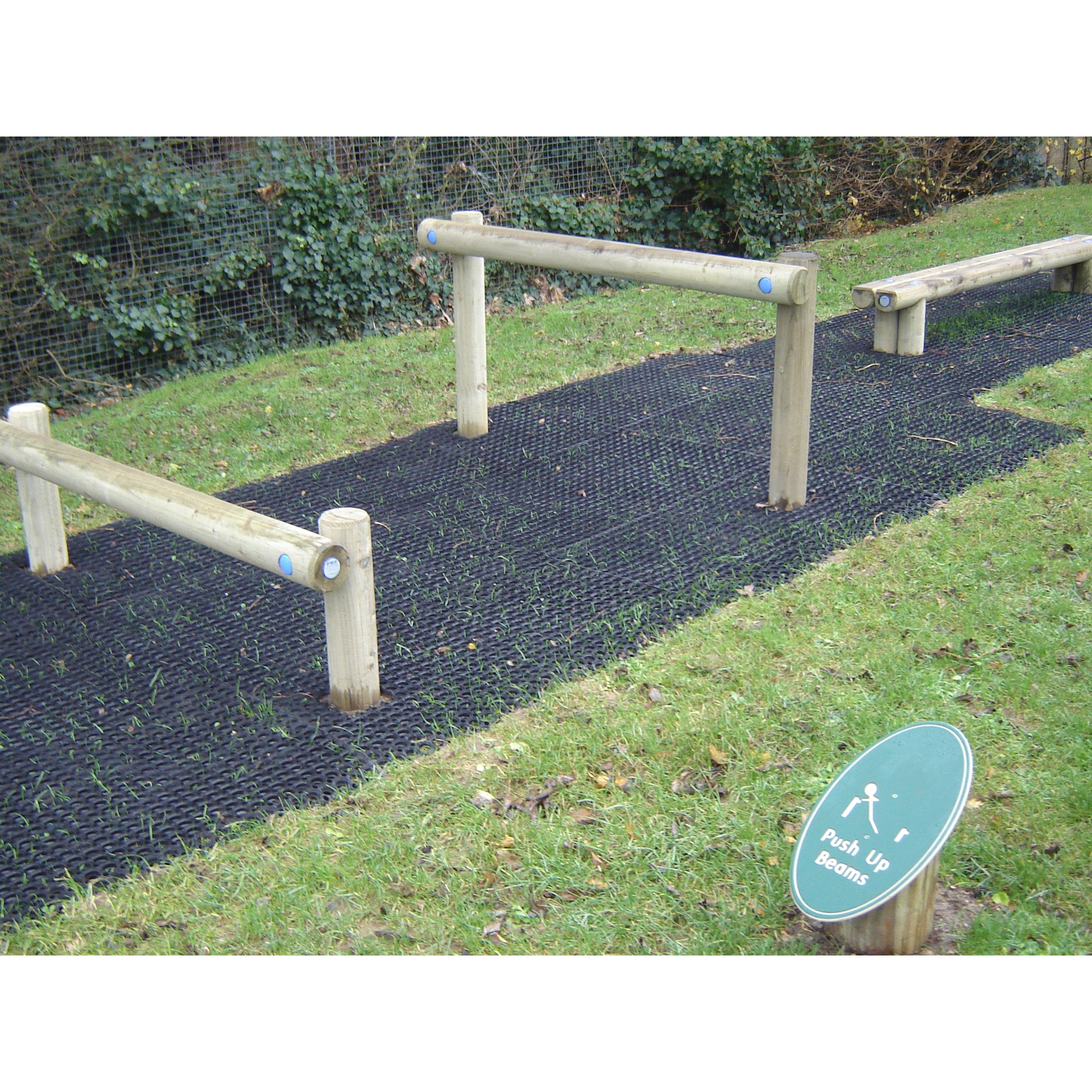 Outdoor fitness area with wooden press-up bars on a black rubber mat, next to a green sign labelled Press-Up Beams.