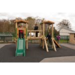Four children play on a wooden playground structure with slides and climbing features, set on a black rubber surface outdoors.
