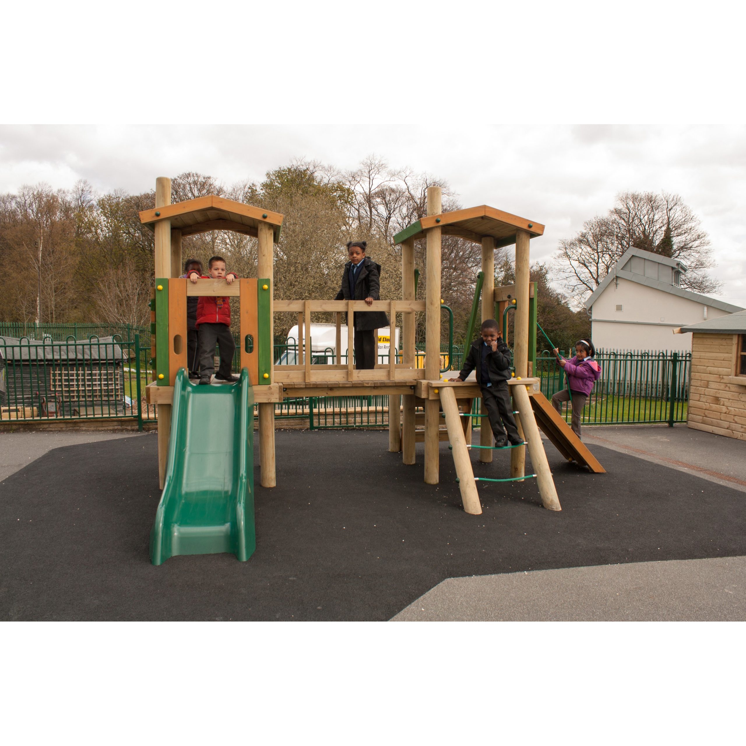 Four children play on a wooden playground structure with slides and climbing features, set on a black rubber surface outdoors.
