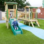 A young child wearing a blue jacket slides down a green slide attached to a wooden climbing frame on artificial grass.