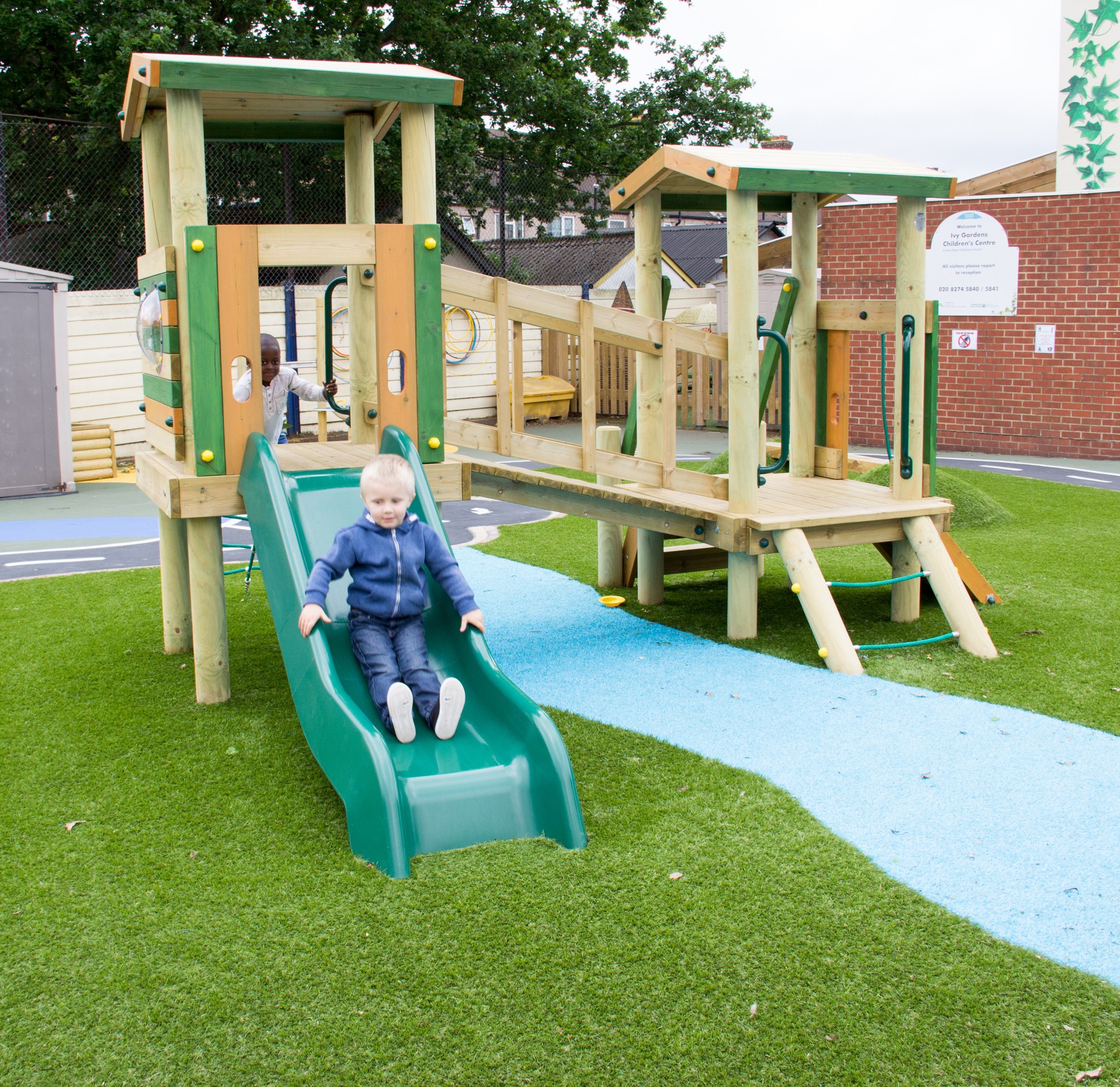A young child wearing a blue jacket slides down a green slide attached to a wooden climbing frame on artificial grass.