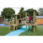 Several children play on a wooden playground structure with ladders, ramps, and a bridge on an artificial grass and rubber surface outdoors.