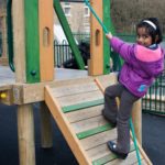 A young girl in a purple jacket climbs a slanted wooden ramp with a rope on a playground structure, looking back and smiling at the camera.