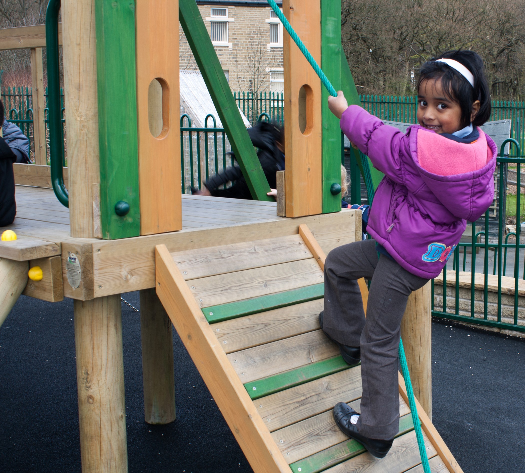 A young girl in a purple jacket climbs a slanted wooden ramp with a rope on a playground structure, looking back and smiling at the camera.