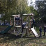 Several children in school uniforms play on a wooden playground structure with slides and climbing features in an outdoor park area surrounded by trees.