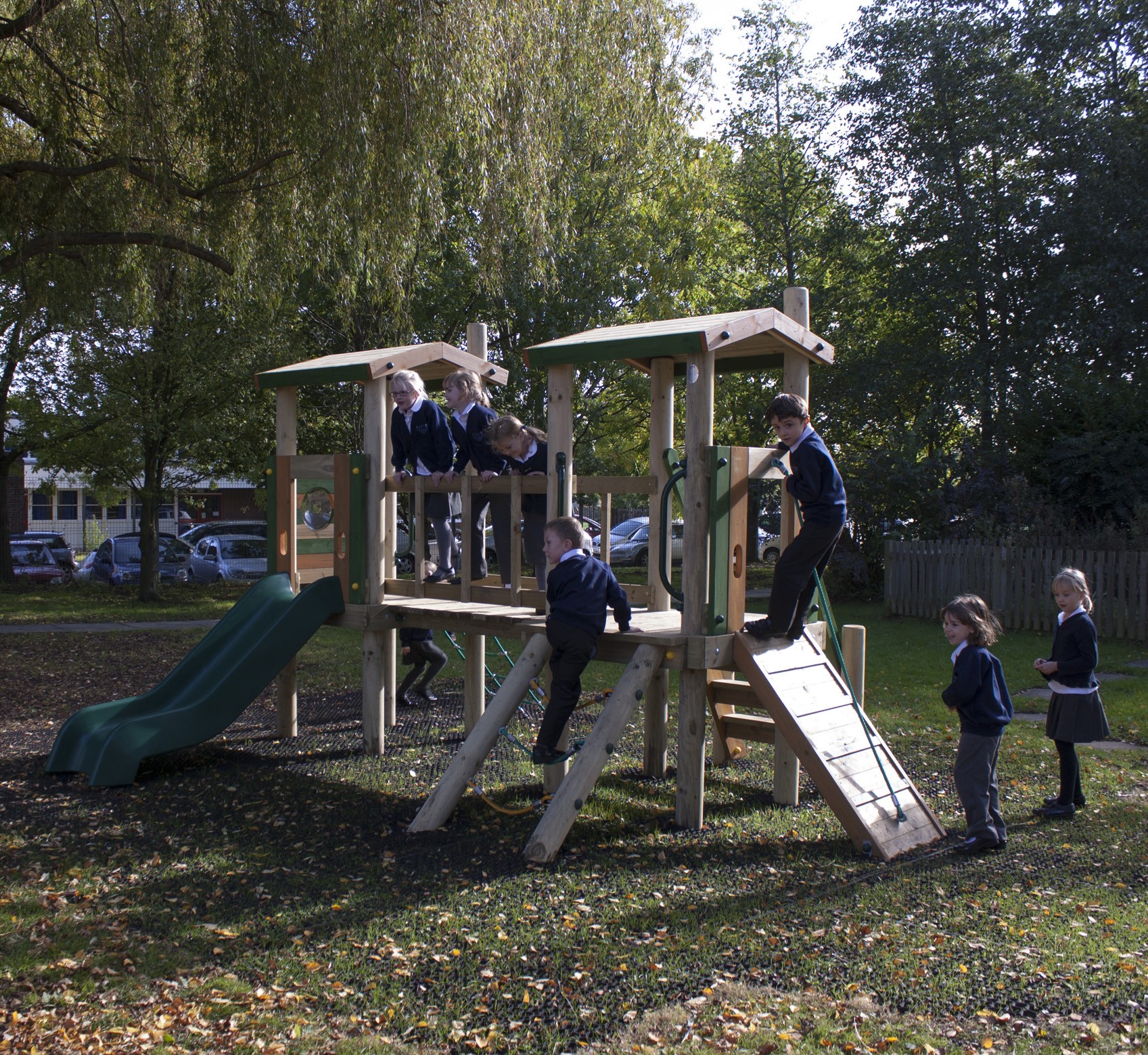 Several children in school uniforms play on a wooden playground structure with slides and climbing features in an outdoor park area surrounded by trees.