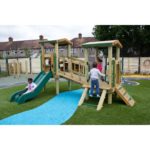 Four children play on a wooden playground structure with a slide, bridge, and climbing features on artificial grass near residential houses.