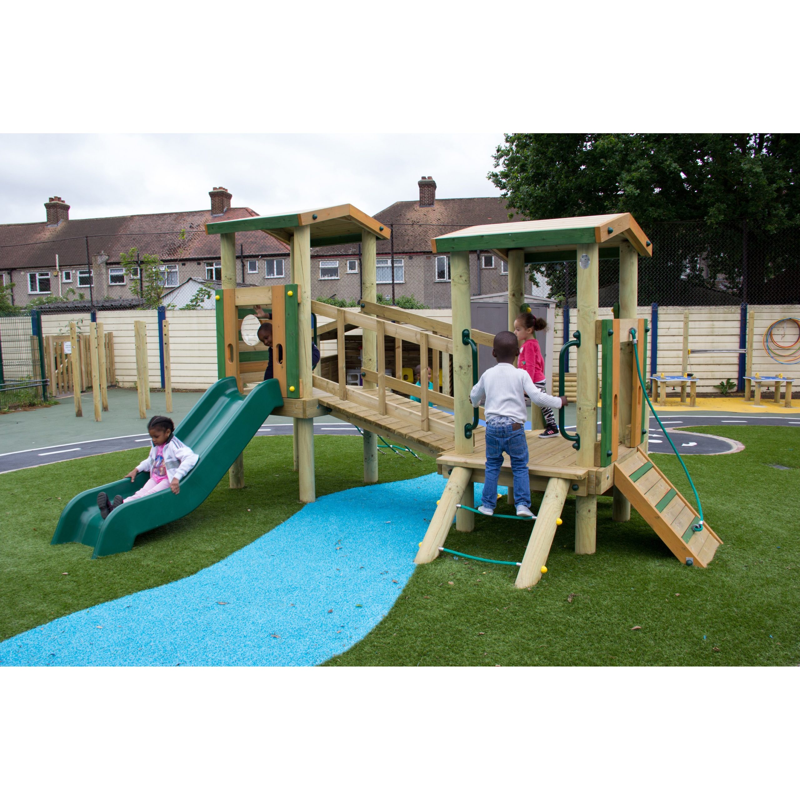 Four children play on a wooden playground structure with a slide, bridge, and climbing features on artificial grass near residential houses.