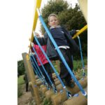 Two children in school uniforms walk across a rope bridge on a playground structure outdoors, with trees in the background.