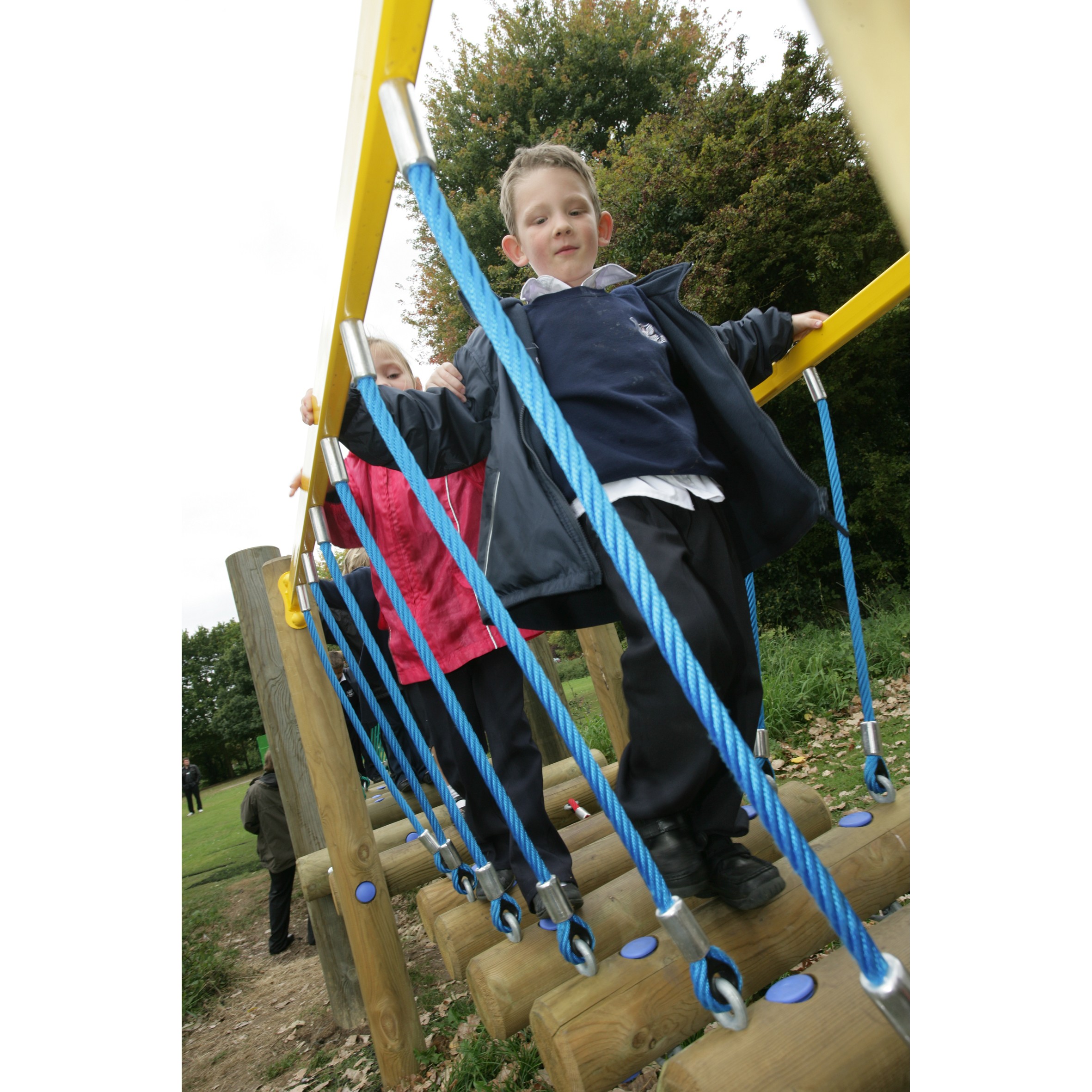 Two children in school uniforms walk across a rope bridge on a playground structure outdoors, with trees in the background.