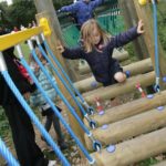 Children are playing on a wooden and rope assault course outdoors. One girl in a navy jacket carefully steps across suspended logs.