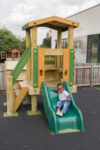 A young explorer enjoys the thrill of sliding down a green Young Explorer - Crusoe 900mm slide attached to a wooden play structure on a playground.