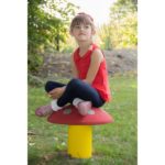 A young girl in a red top and blue leggings sits cross-legged on a mushroom-shaped seat outdoors in a grassy area.