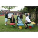 A group of children in school uniforms sit on mushroom-shaped stools outdoors while one child draws on a whiteboard.