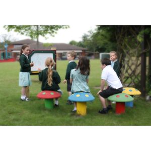 A group of children in school uniforms sit on mushroom-shaped stools outdoors while one child draws on a whiteboard.