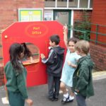 Four children play at a red pretend post office stand outdoors; one stands behind the counter while others interact in front.
