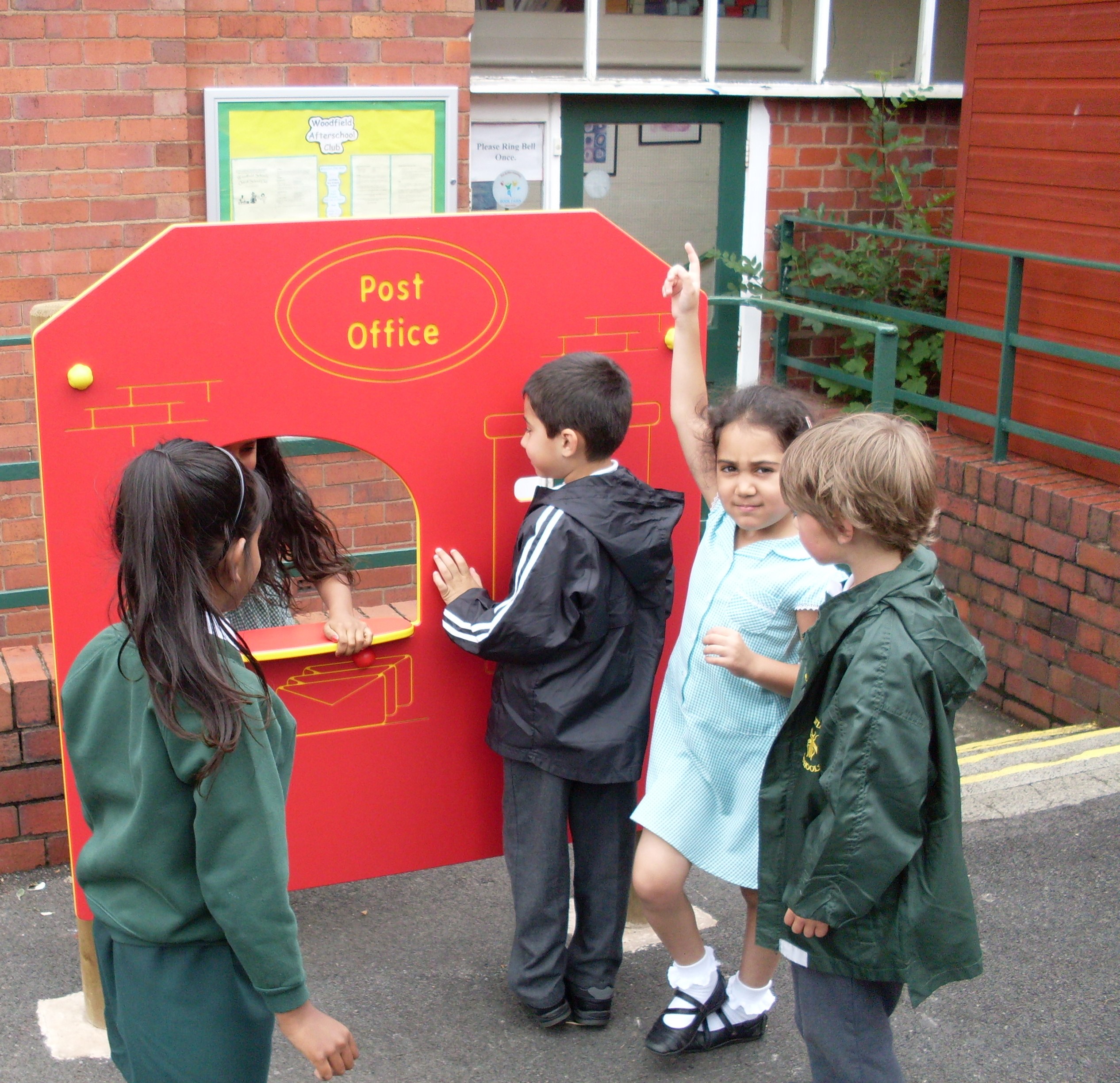 Four children play at a red pretend post office stand outdoors; one stands behind the counter while others interact in front.