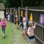 Several young children walk and play along a wooden fence decorated with large, colourful numbers outdoors on a cloudy day.
