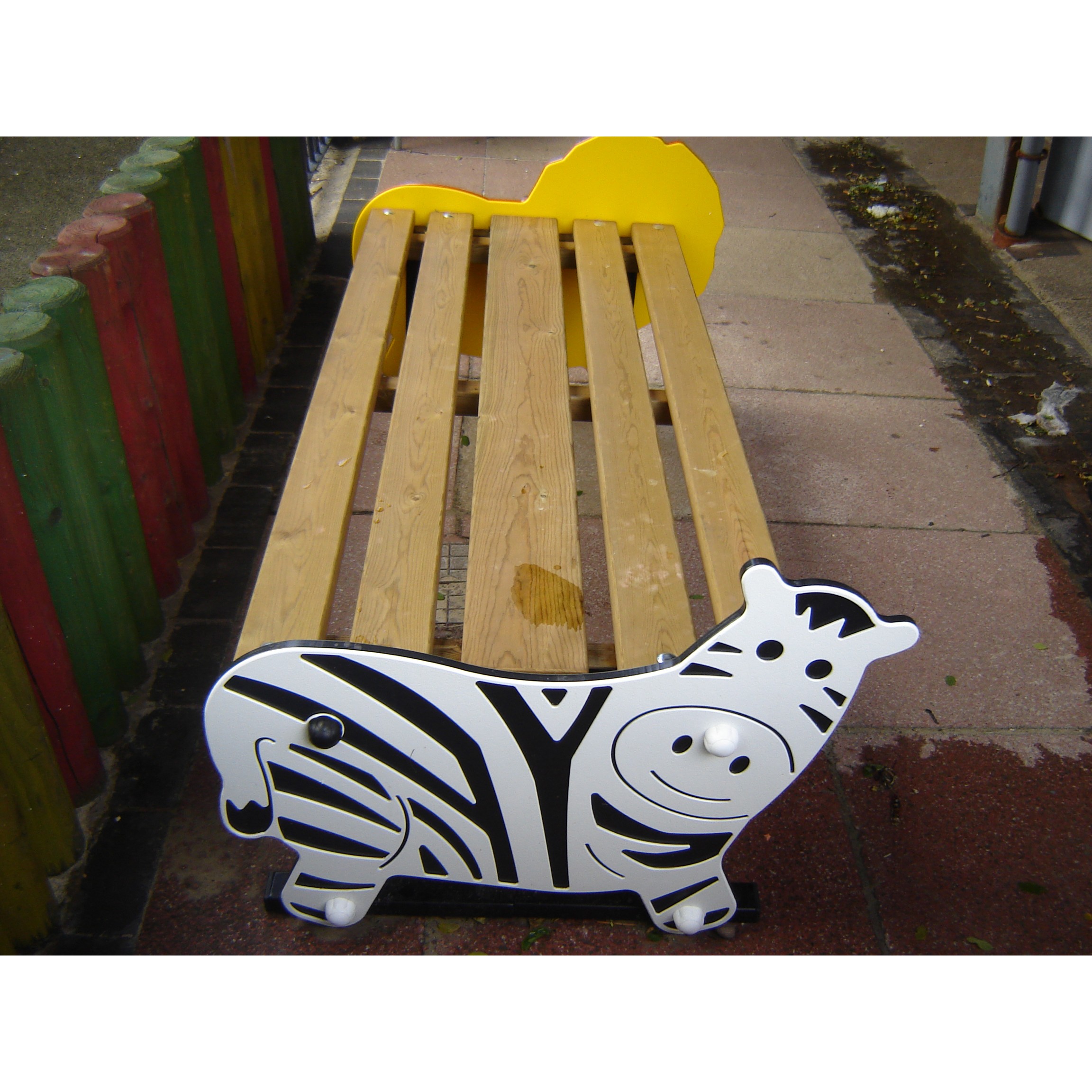 A wooden bench with ends shaped and painted like a zebra, featuring a yellow lion figure at the opposite end, placed on a playground area.