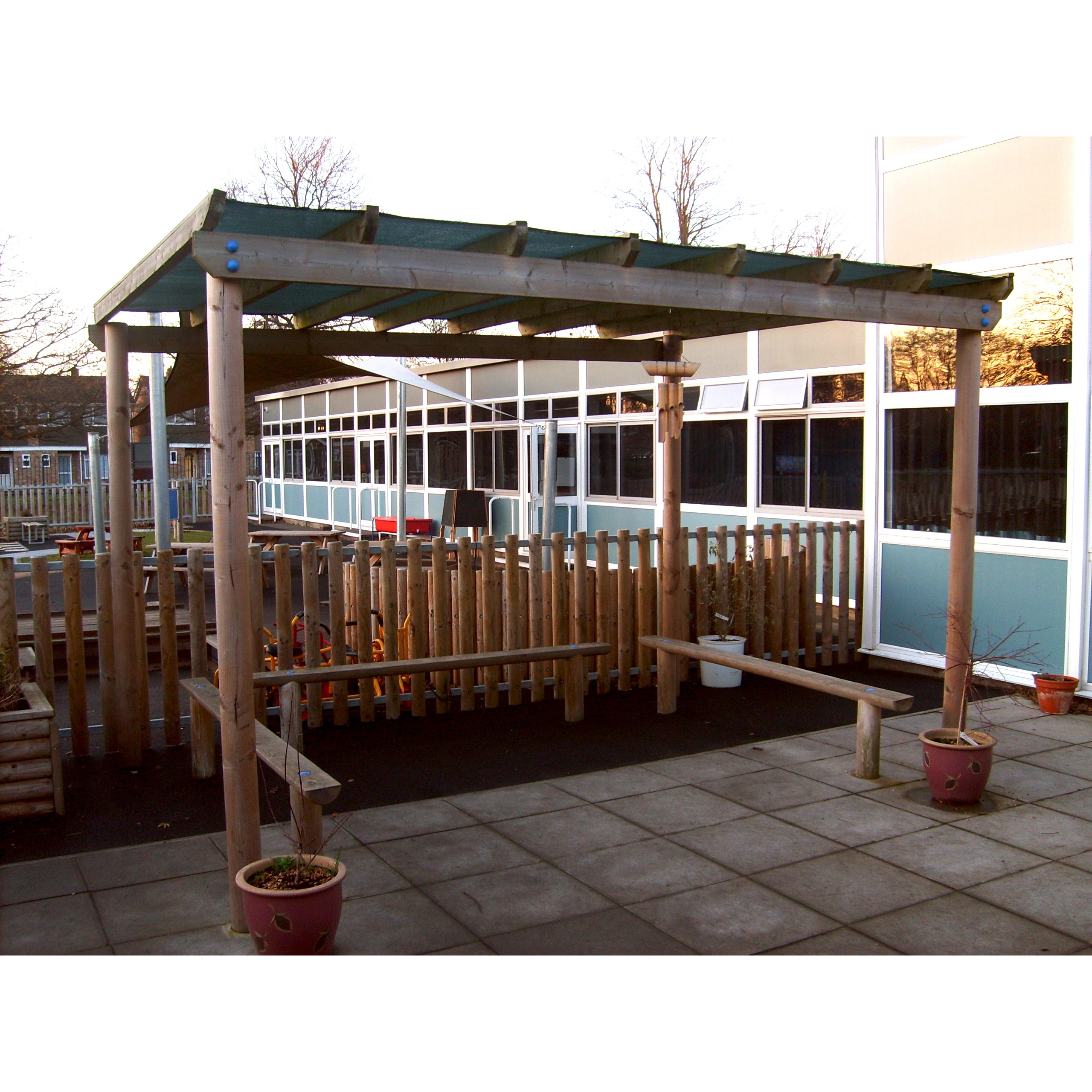 A wooden pergola with a bench stands on a paved area near a fenced garden outside a modern building with large windows.