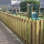 A fenced outdoor play area with a wooden climbing frame and a green slide, surrounded by grass and a paved pathway.