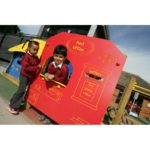Two young children in school uniforms play at a red climbing frame designed to look like a post office in an outdoor playground.