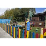 Outdoor playground area with colourful fence, play equipment, shade canopy, and informational sign, adjacent to a brick building and surrounded by trees.