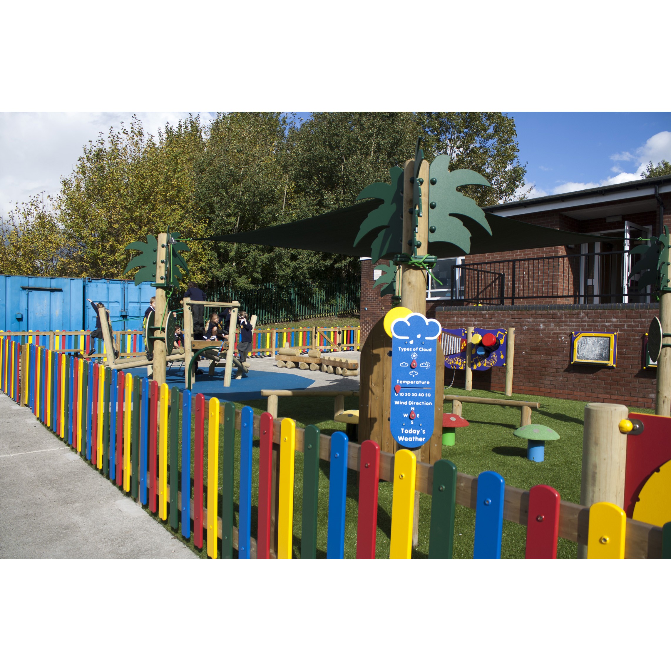Outdoor playground area with colourful fence, play equipment, shade canopy, and informational sign, adjacent to a brick building and surrounded by trees.