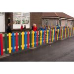 Children in coats walk along a playground behind a colourful wooden fence outside a brick building with decorative window art.