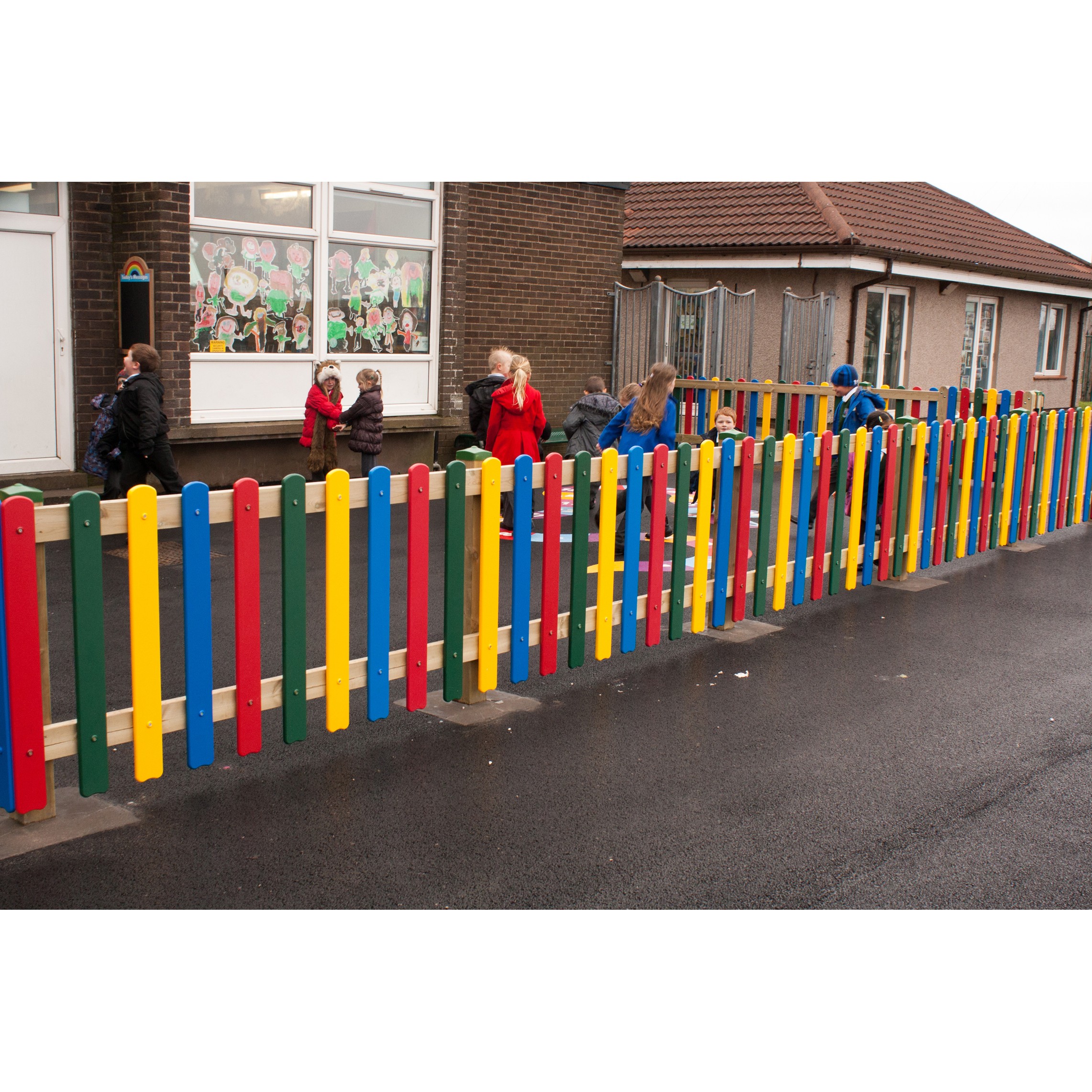Children in coats walk along a playground behind a colourful wooden fence outside a brick building with decorative window art.