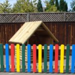 A small wooden playhouse stands behind a colourful fence with red, yellow, blue, and green vertical slats, in front of a tall wooden fence and trees.