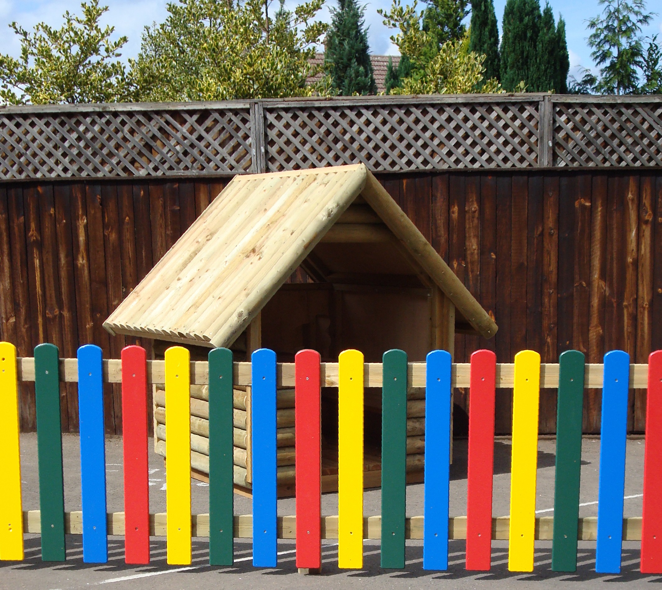 A small wooden playhouse stands behind a colourful fence with red, yellow, blue, and green vertical slats, in front of a tall wooden fence and trees.