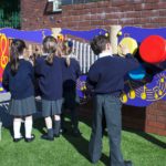 Four children in school uniforms play with a colourful outdoor musical wall featuring various percussion instruments mounted against a brick wall.
