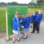 Six young children in school uniforms interact with a green play panel labelled Garage on a playground.