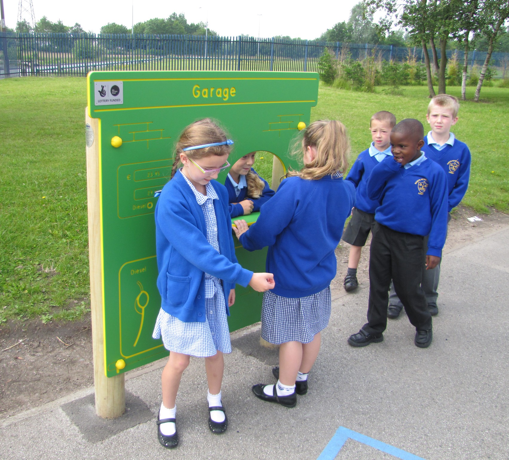 Six young children in school uniforms interact with a green play panel labelled Garage on a playground.