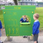 Three children in school uniforms play at an outdoor pretend garage station with labelled fuel pumps and a window. Two children face each other across the window.