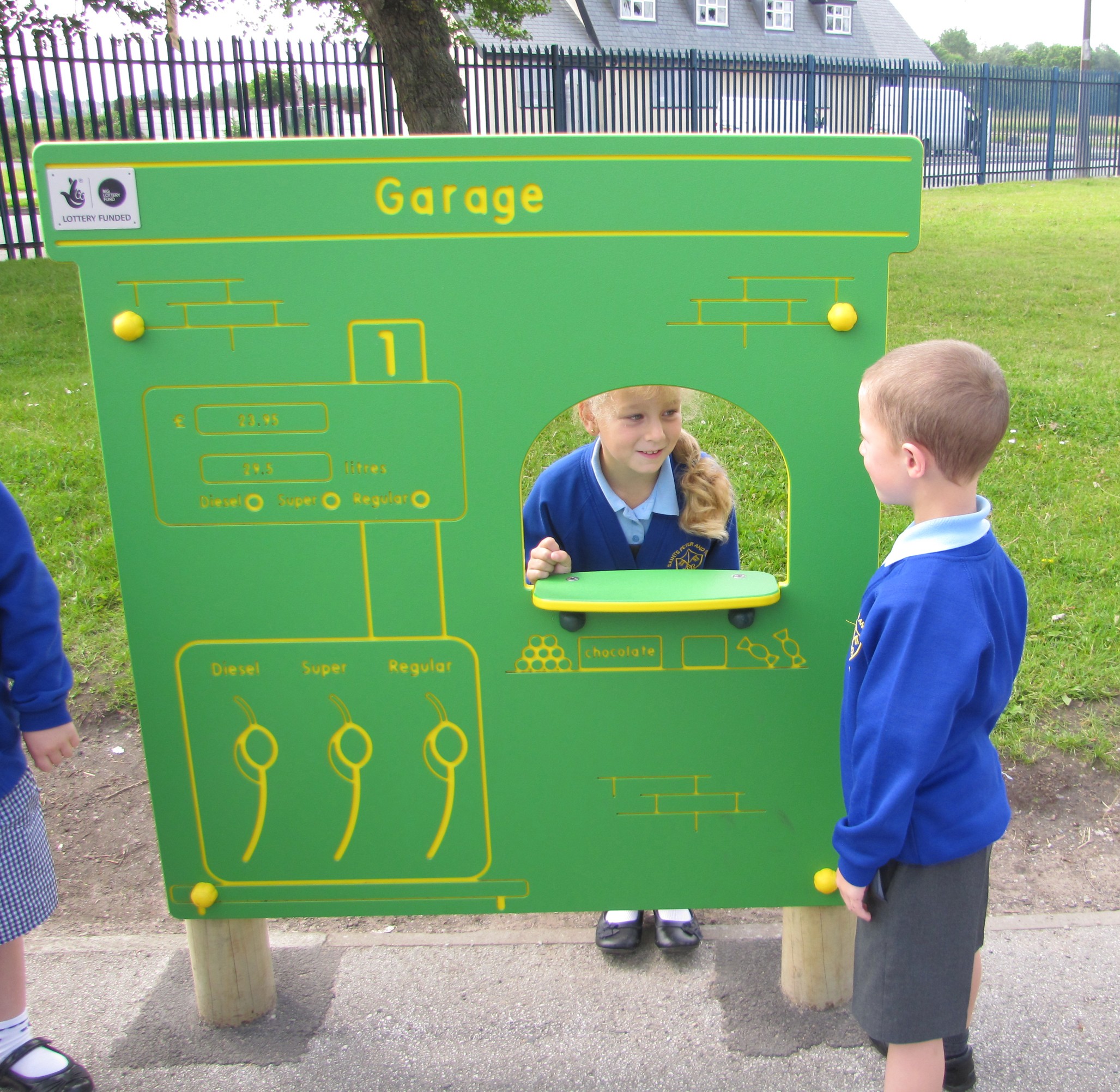 Three children in school uniforms play at an outdoor pretend garage station with labelled fuel pumps and a window. Two children face each other across the window.