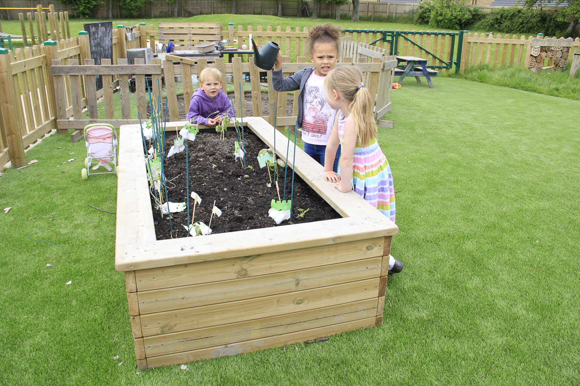 Three young children are tending to plants in a 55cm Log Planter on a grassy playground; one child is carefully watering the thriving garden bed.