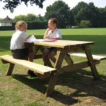 Two children wearing school uniforms sit at a wooden picnic table on a grassy field, talking and looking at papers and exercise books on the table. Trees and a house are in the background.