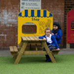 Two young girls in school uniforms sit and stand by a wooden picnic table in a playground area with play shop panels in the background.