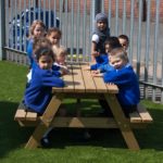 A group of young children in blue school uniforms sit around a wooden picnic table on artificial grass, outdoors near a metal fence.