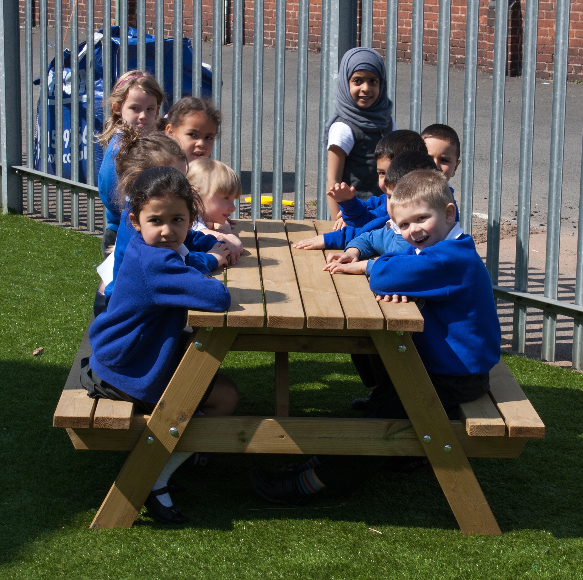 A group of young children in blue school uniforms sit around a wooden picnic table on artificial grass, outdoors near a metal fence.