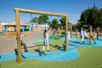 Several children in school uniforms play on a climbing frame with ropes on a school playground under a clear blue sky.