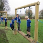 Six young children in blue uniforms balance and play on a wooden outdoor playground structure with ropes, set on grass near trees, engaging in outdoor maths activities using playground equipment.