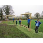 Four children in blue jackets play on a wooden balance and climbing structure in an outdoor grassy area with trees in the background.