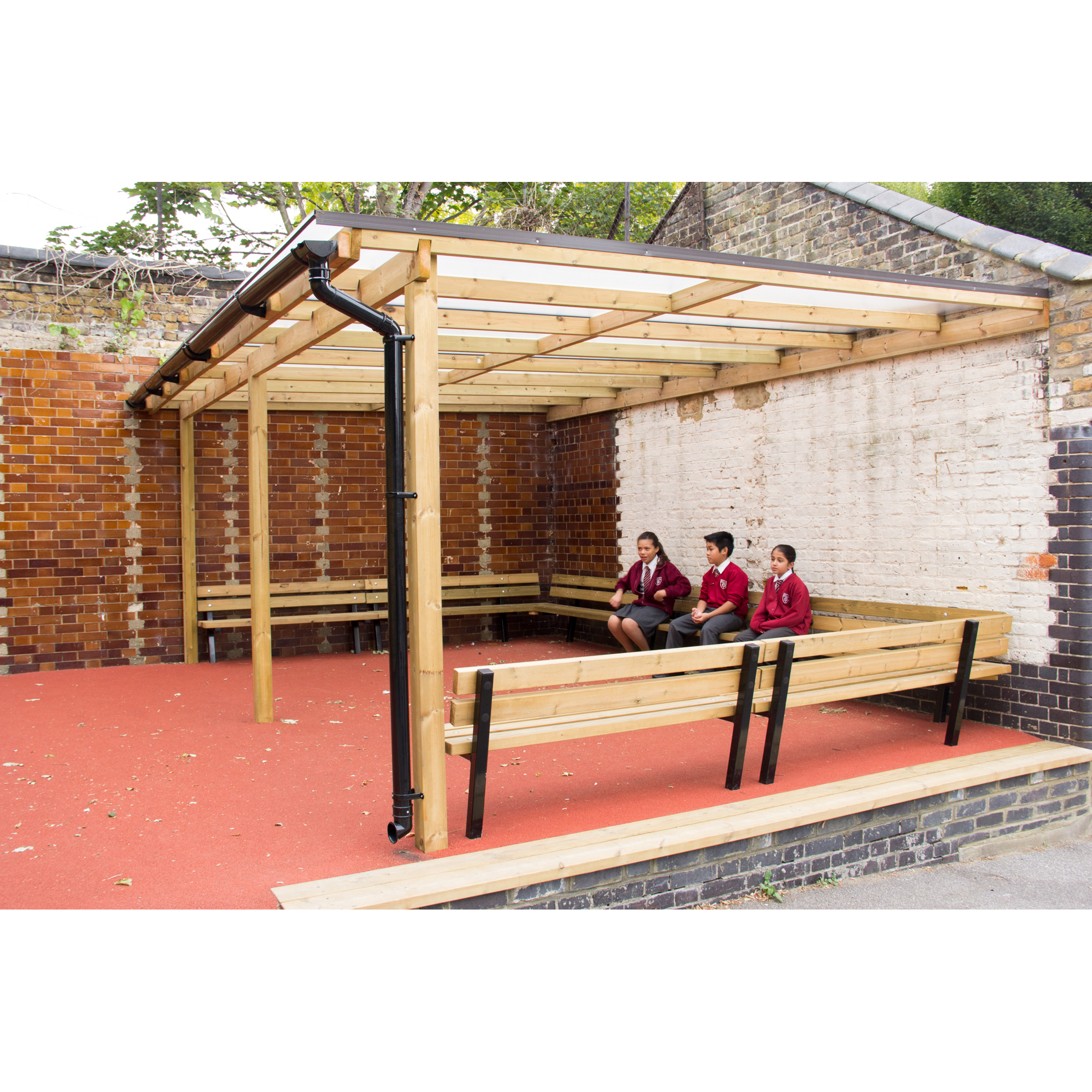 Three children in school uniforms sit on a bench under a wooden shelter in a corner of a yard with brick walls and a red floor.